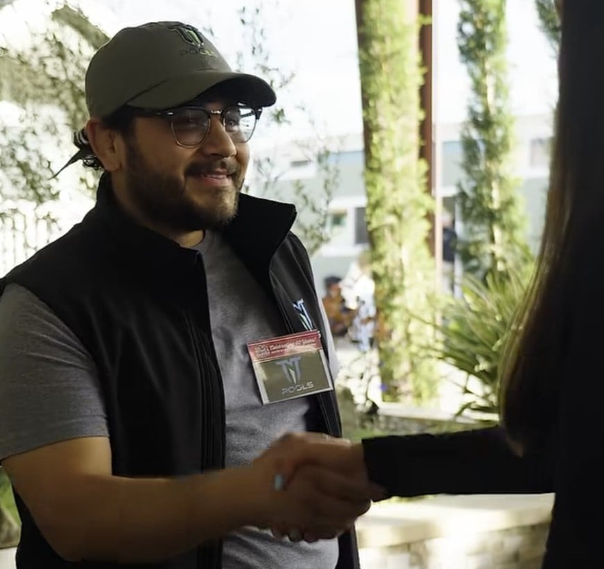 Man wearing olive cap and glasses handshaking with another person indoors near greenery and windows